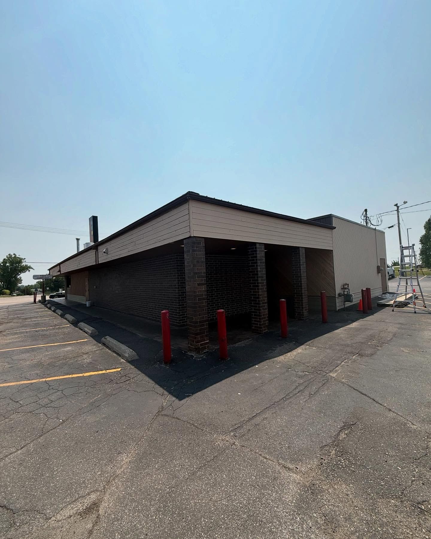 Abandoned brick building with dark front, red poles, and a paved parking lot on a sunny day.