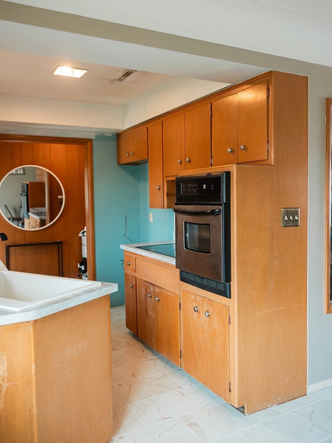 Mid-century kitchen with light wood cabinets, a built-in oven, and a turquoise wall. A circular mirror is visible.