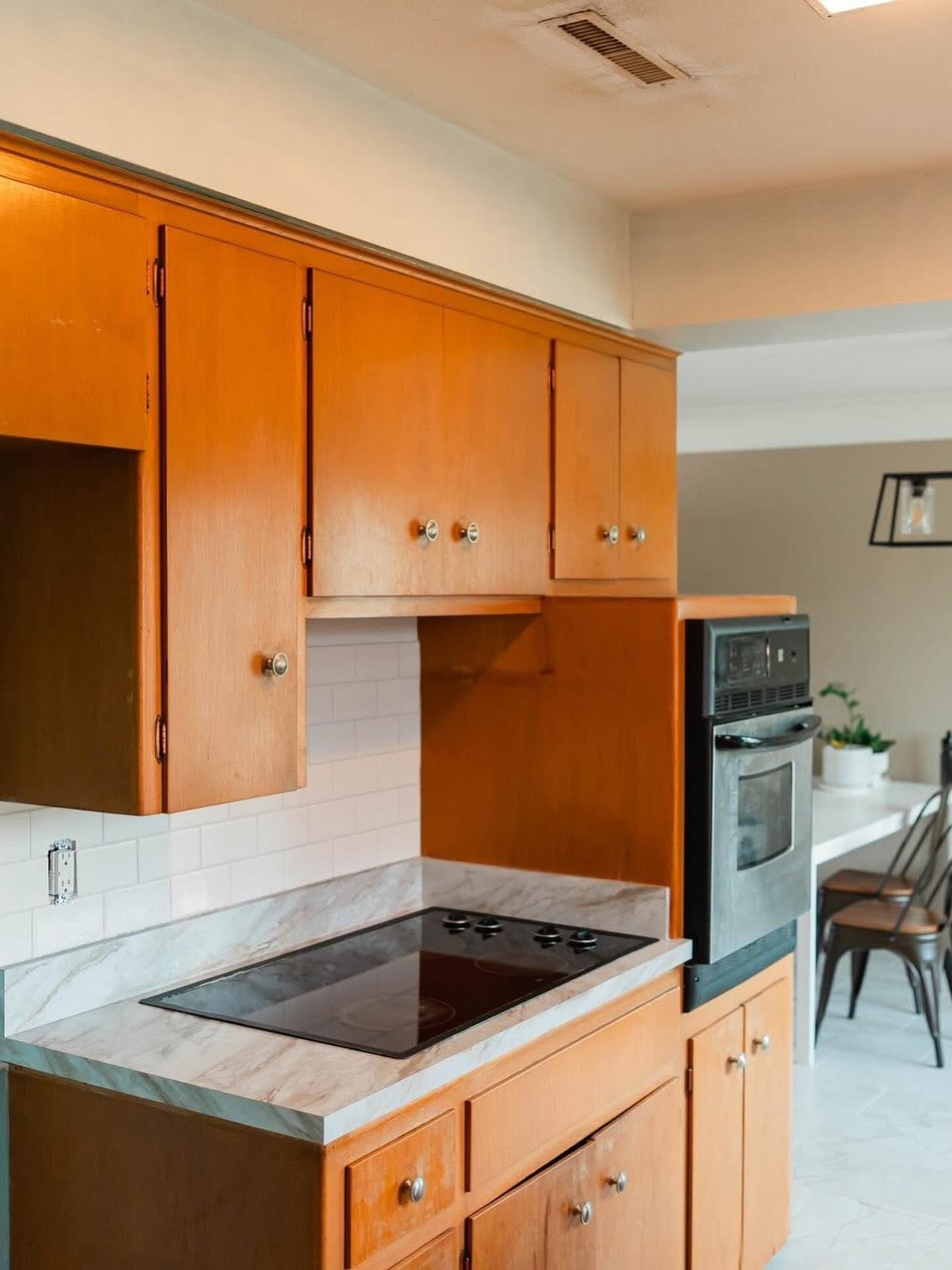 Kitchen with light wood cabinets, stovetop, built-in oven, and a dining area in the background.