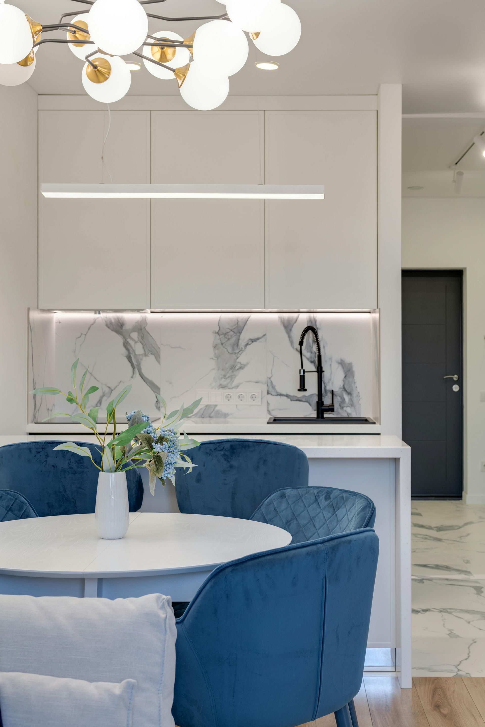Modern kitchen with white cabinets, marble backsplash, blue chairs, and a round table.