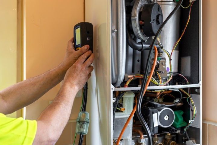 Technician using a handheld meter to inspect an open furnace control panel with exposed wiring