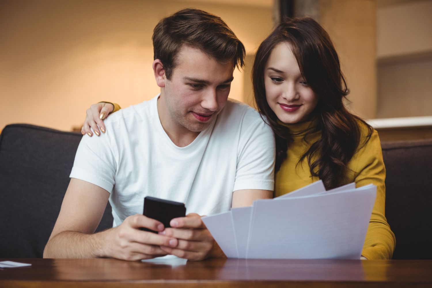 A man and a woman are sitting at a table looking at papers and a cell phone.