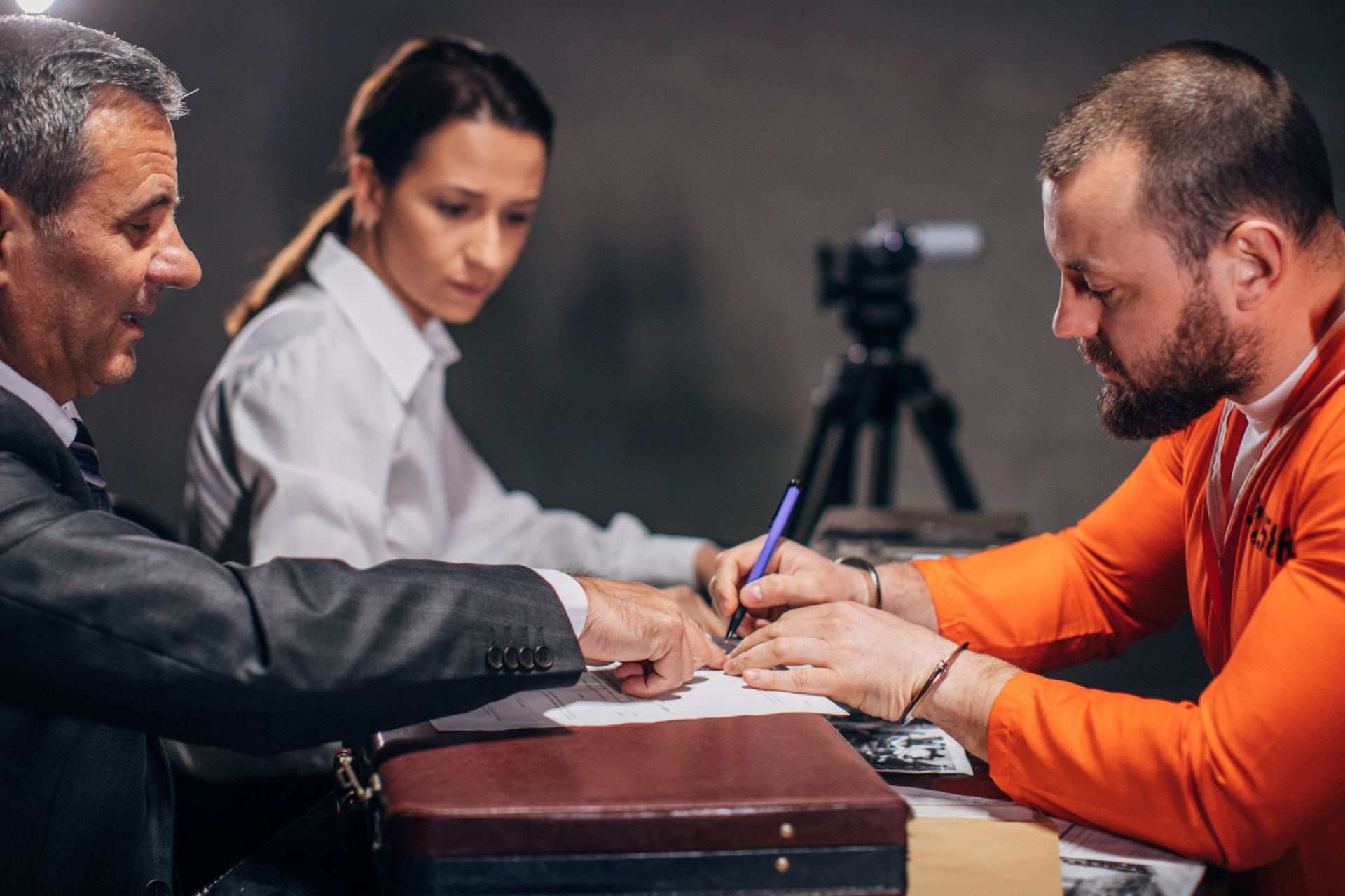 A man in an orange jumpsuit is signing a document while sitting at a table with two people.