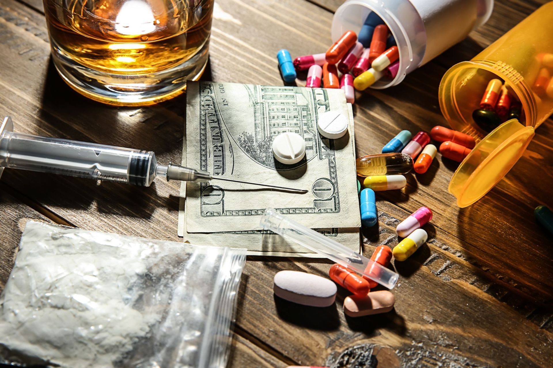 A wooden table topped with pills , money , a syringe , and a glass of whiskey.
