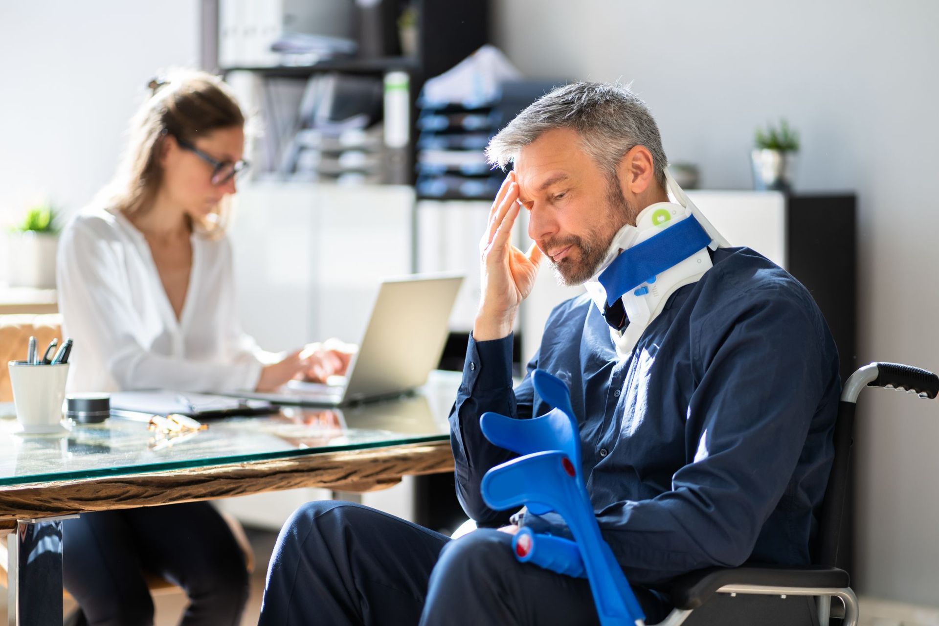 A man in a wheelchair with a neck brace and crutches is sitting at a desk.