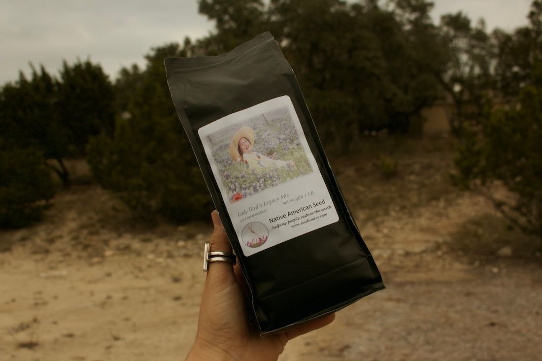 Hand holding black seed bag with white label that says Native American Seed; outdoor setting.
