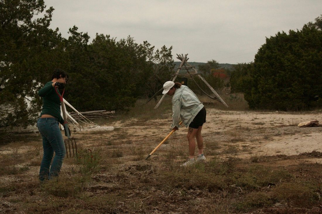 Two people raking a dirt field. Trees in the background, overcast sky.