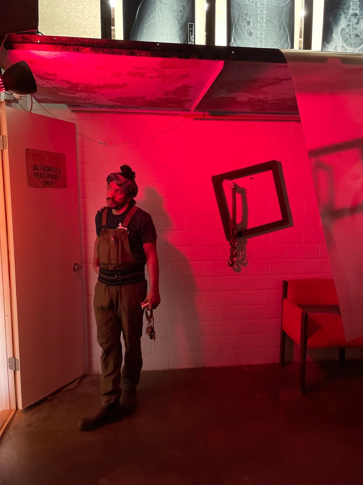 Carlos Castaneira, founder of the Arts and Culture Lab in Houston, Texas, in overalls stands in a red-lit room with a doorway, a chair, and a framed minimalist sculpture on the wall.