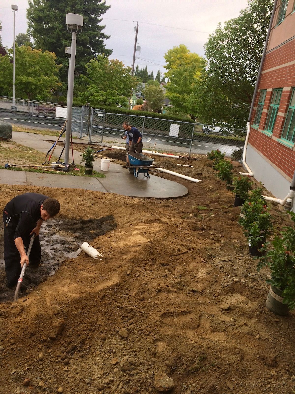 Two workers landscaping: one shoveling dirt, the other using a wheelbarrow, next to a brick building.
