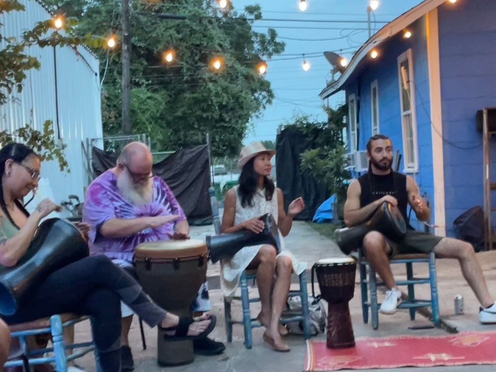 Group of people playing drums outdoors at dusk, under string lights, at the first artist residency site of the Arts and Culture Lab in Houston, Texas.