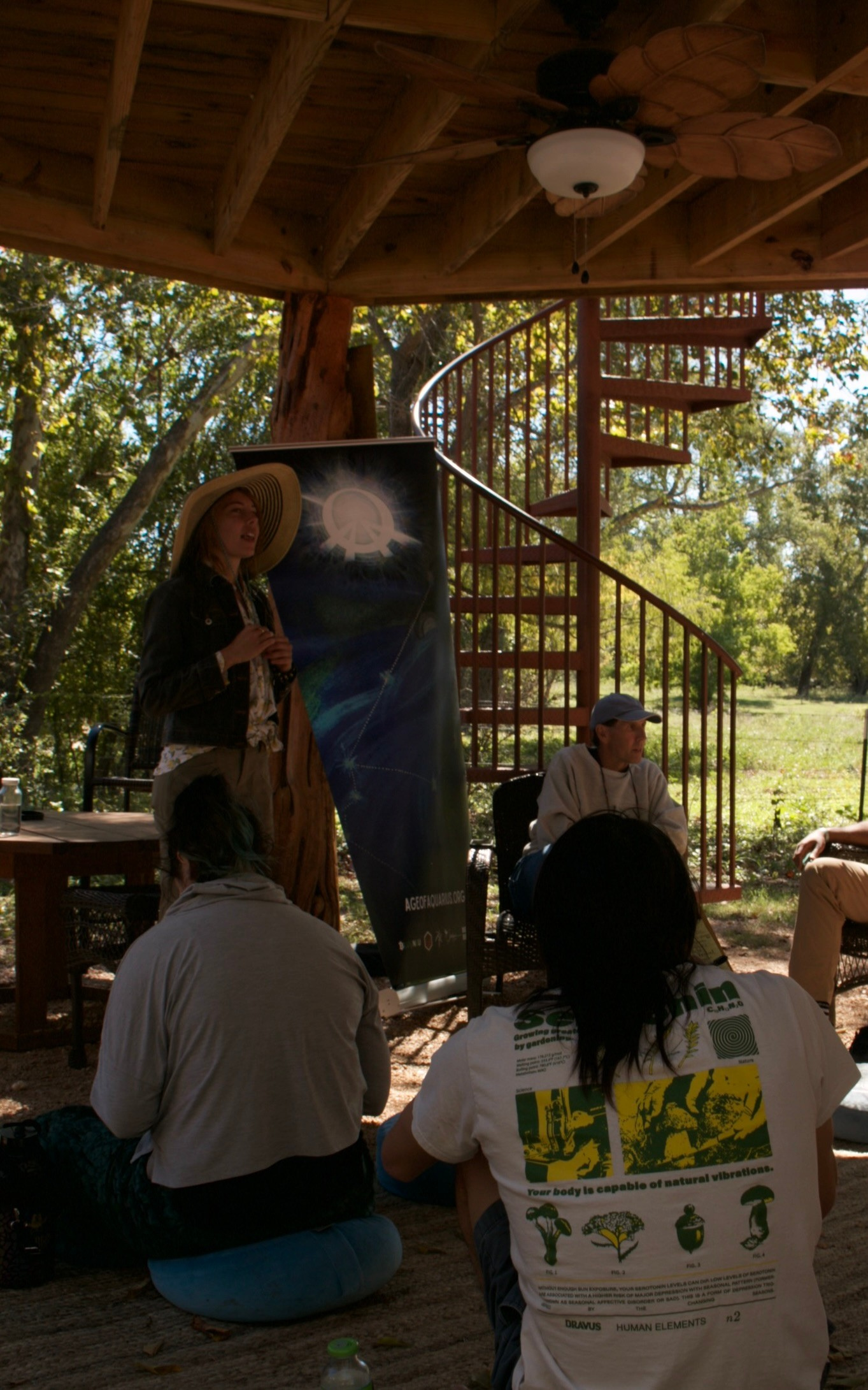Permaculture teacher, Marlo gives a presentation under a wooden pergola with audience. Features a poster of the solar eclipse coinciding with the event.