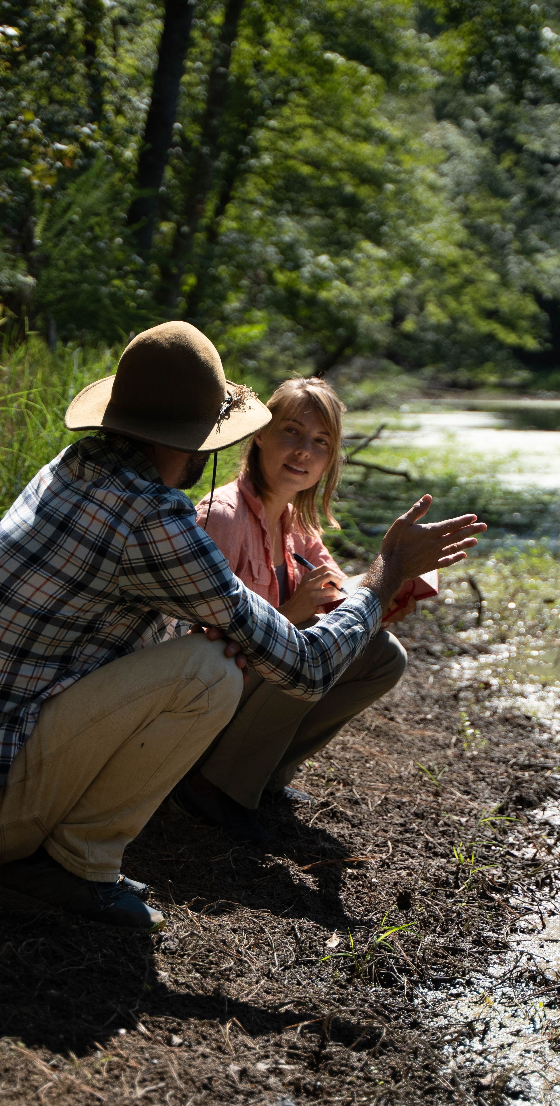 Two people squatting near water, one pointing, sunny setting.