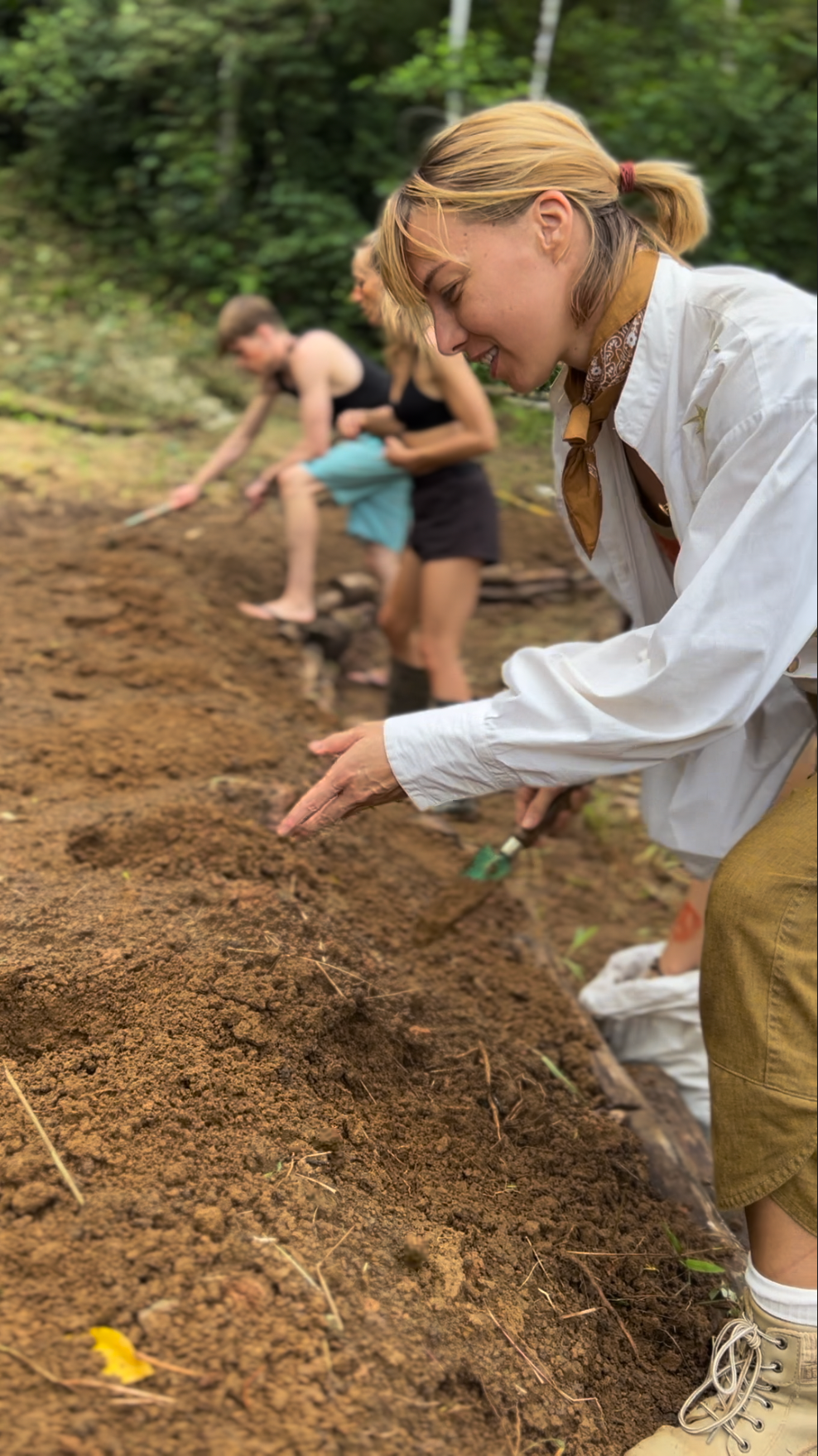 Marlo Weekley planting root stock in a syntropic agroforestry garden bed, with two others behind her. Outdoors, sunny. Brown clay soil.