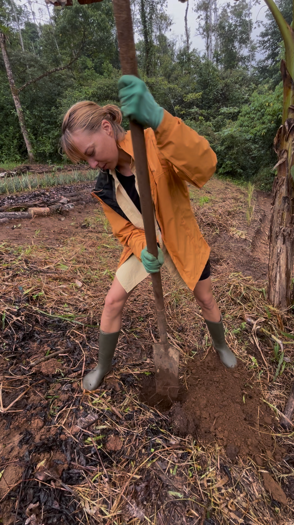 Marlo Weekley in raincoat and gloves digging in a muddy garden with a long-handled post hole digger.