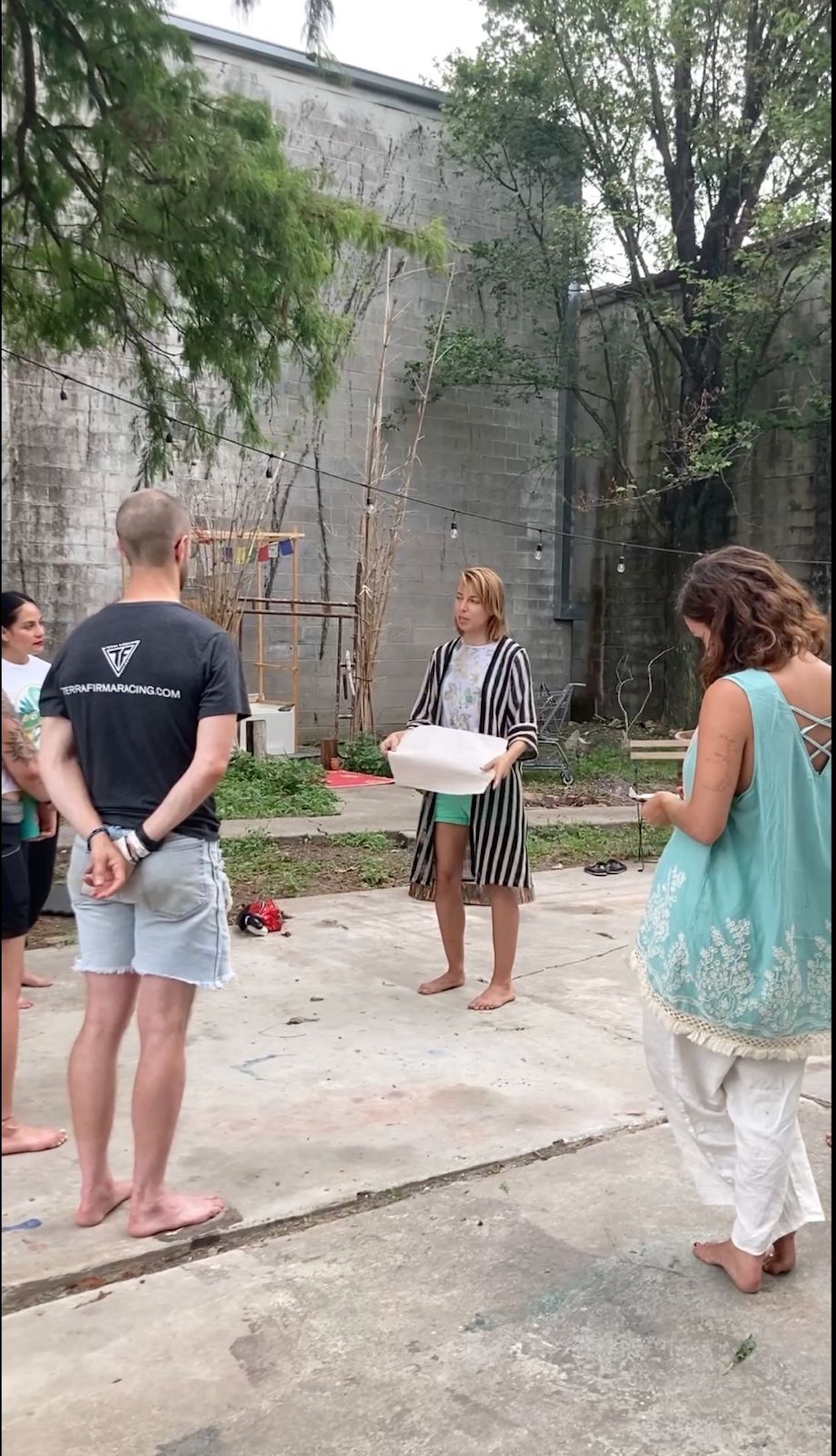 Marlo Blythe teaching a permaculture class at the Arts and Culture Lab Artist Residency, outdoors at the first Urban Permaculture Rainforest Initiative garden site. A few trees and small green space with a weathered brick wall backdrop.