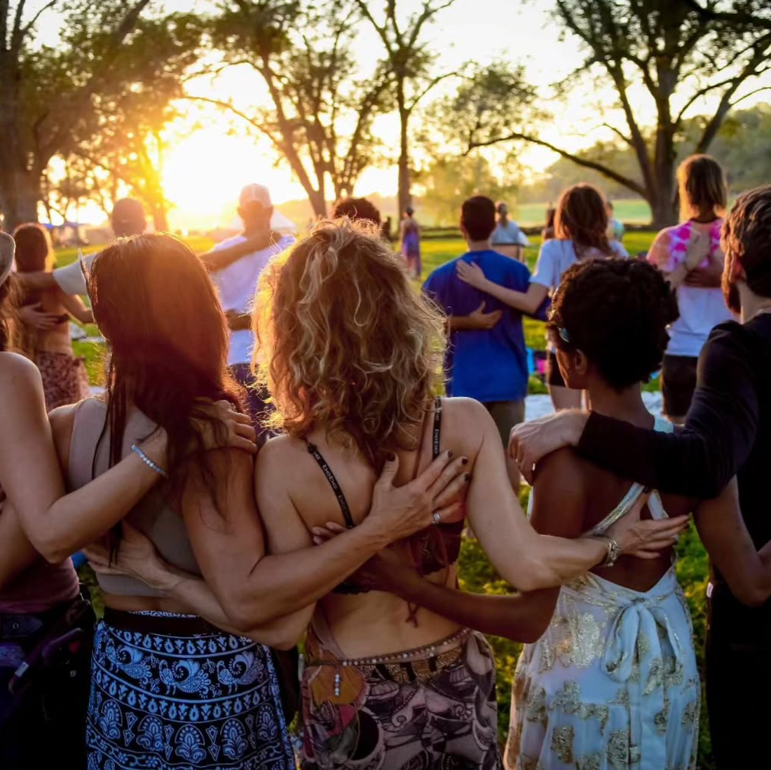 People in a circle with arms around each other, facing a sunset in a grassy field.