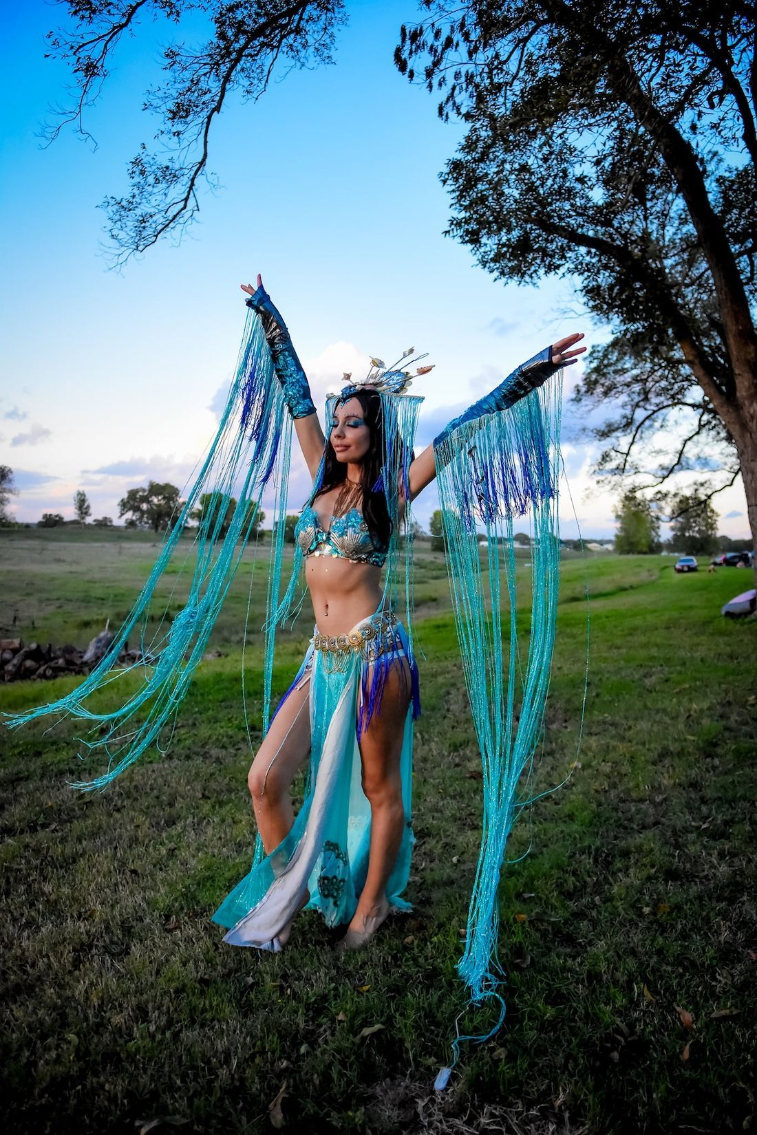 Woman in blue fringe costume with arms raised in a grassy field, under an oak tree.