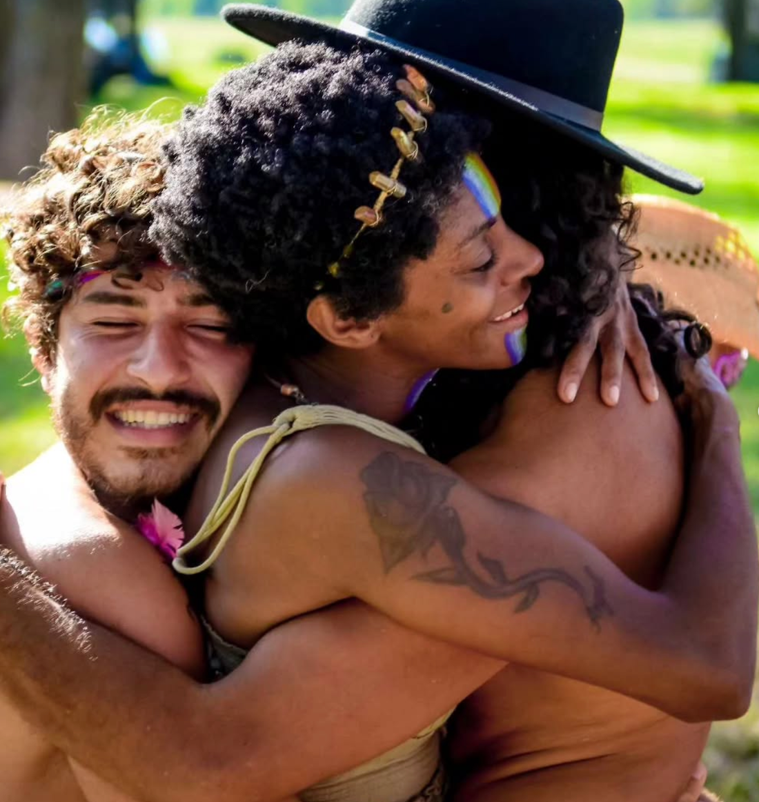 Three people embracing outdoors, smiling. One wears a hat and headband, others have flowers. Rainbow makeup.