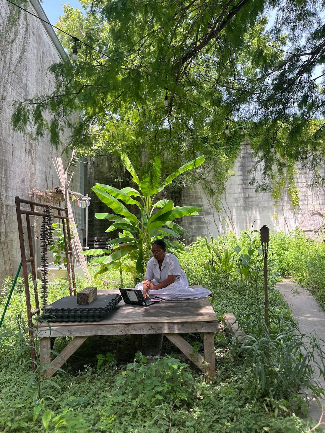 Artist in residence working at a wooden table outdoors, surrounded by plants, under a tree in the Urban Permaculture Rainforest Initiative's first garden.