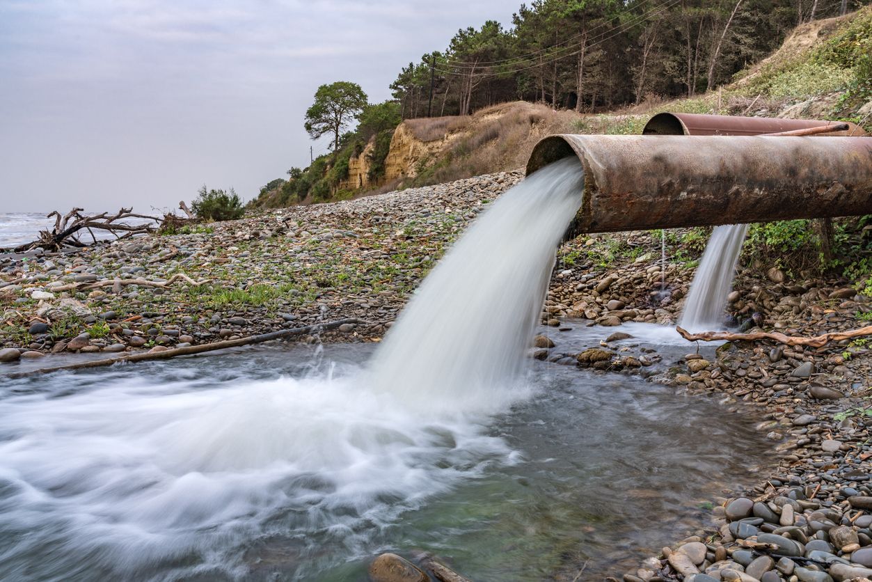 Water gushes from large pipes into a rocky stream near a shoreline, with trees in the background.