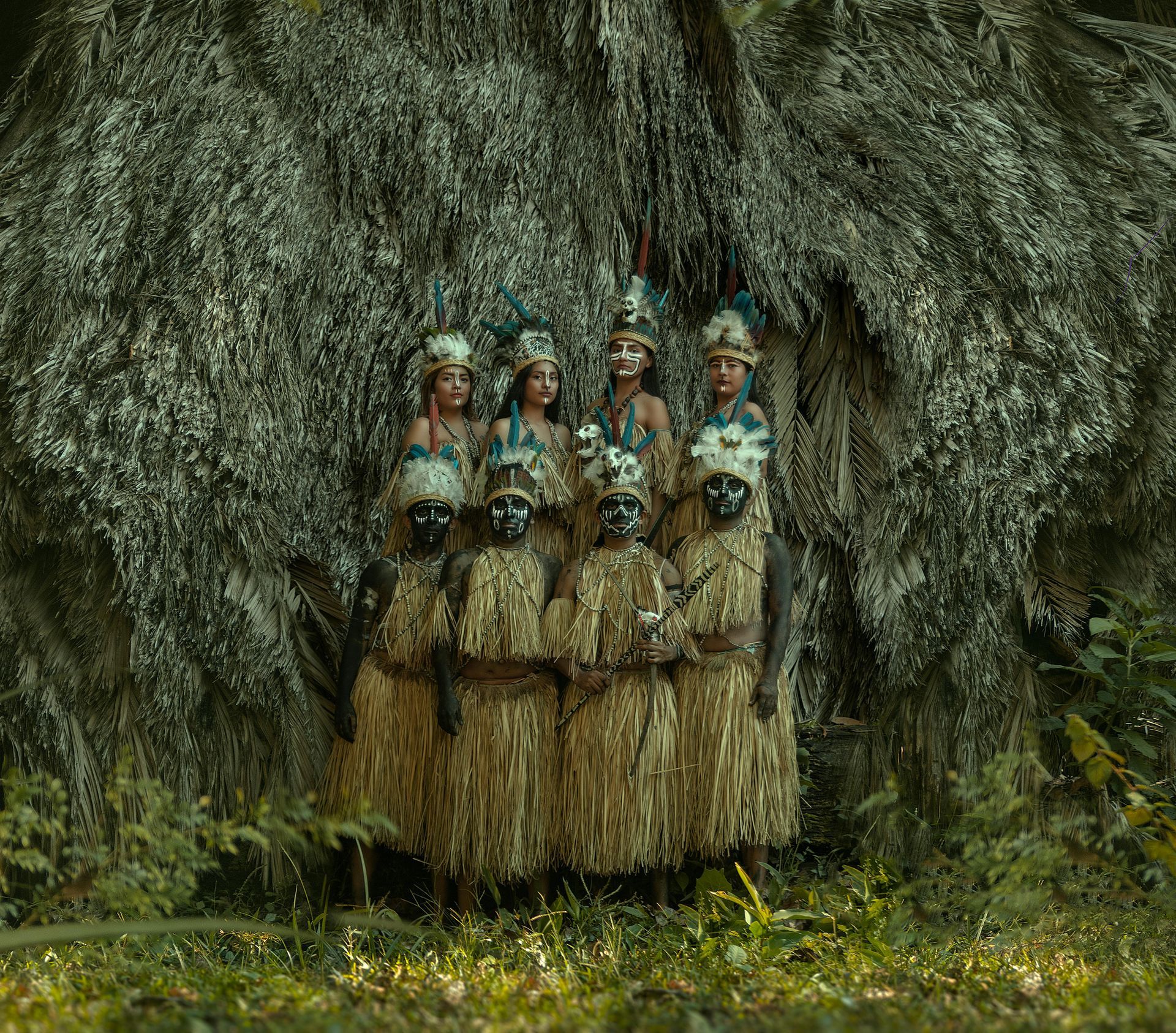 A group of indigenous Brazilian Amazonian people in elaborate costumes and headdresses stand within a natural leafy structure in the background.