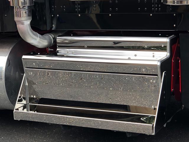 Chrome step box on the back of a semi-truck near a large metal tank and exhaust pipe.