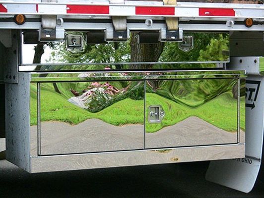 Shiny chrome toolbox on a truck trailer, reflecting a grassy area and trees.