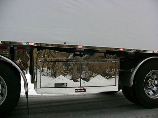 Stainless steel storage box on a semi-trailer, reflecting surroundings, wheels and fenders visible.