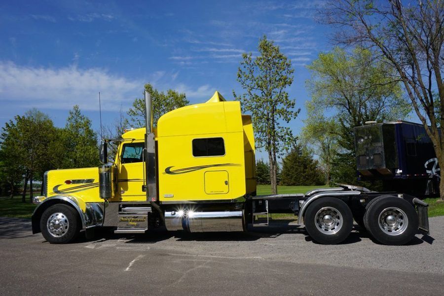 Yellow semi-truck parked on pavement with trees in the background under a blue sky.
