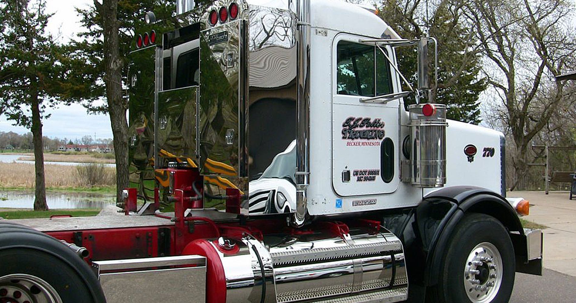 White semi-truck with chrome details, red frame. Park setting, partially visible lake and trees. 