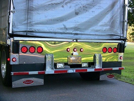 Rear of a semi-truck trailer with chrome accents, red lights, and a license plate, parked outdoors.