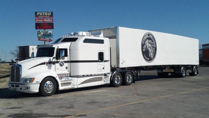 White semi-truck with trailer featuring a logo, parked at a Petro stop under a blue sky.