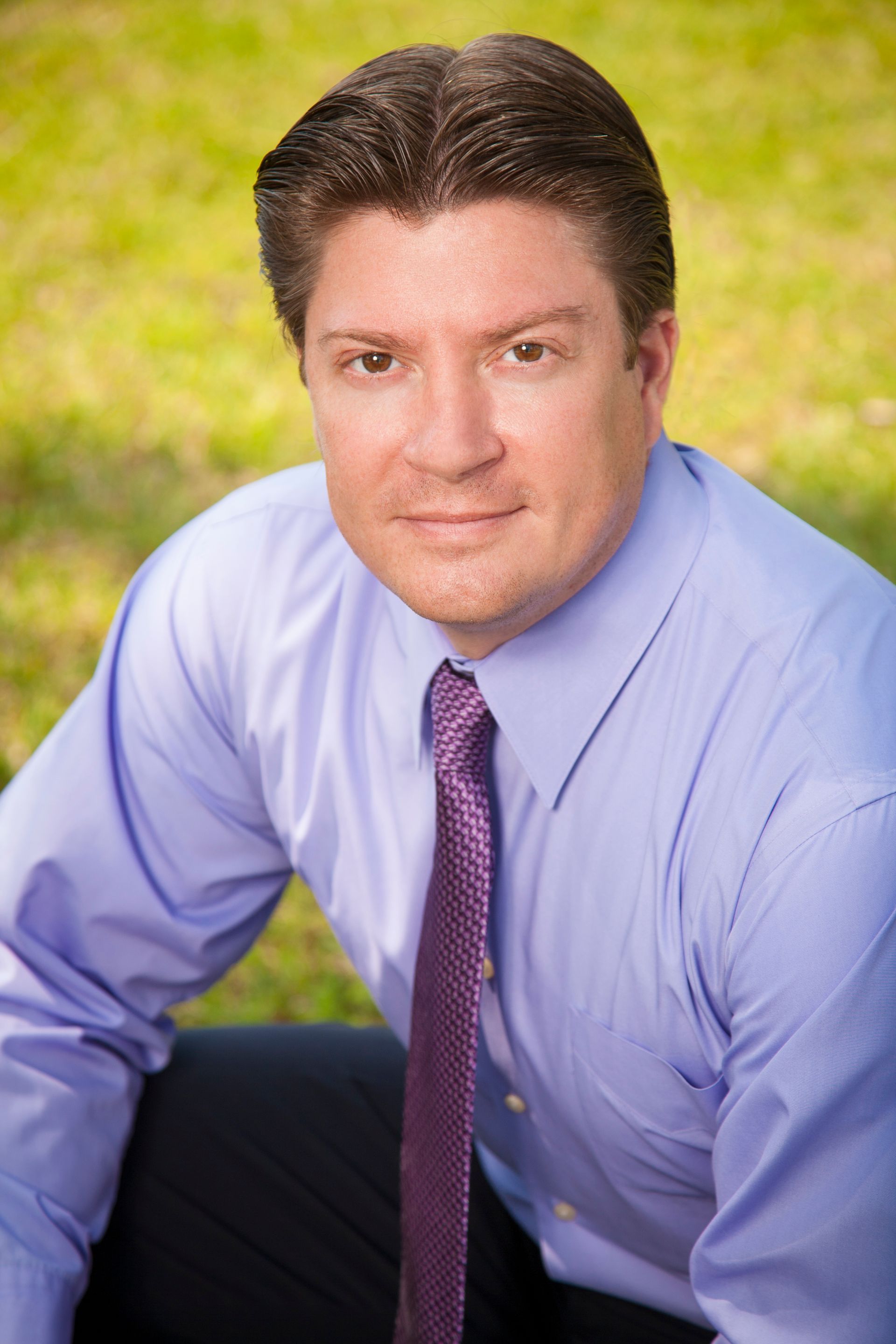 Man in blue shirt and purple tie, smiling, seated outdoors on grass.