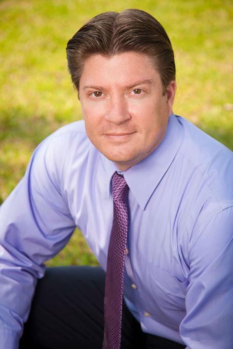 Man in blue shirt and purple tie, kneeling on grass, smiling.