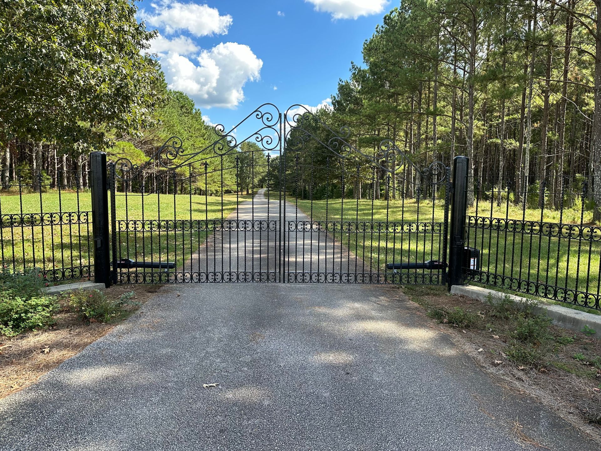 Gravel driveway leading to a black iron gate with a tree-lined path beyond, under a partly cloudy sky
