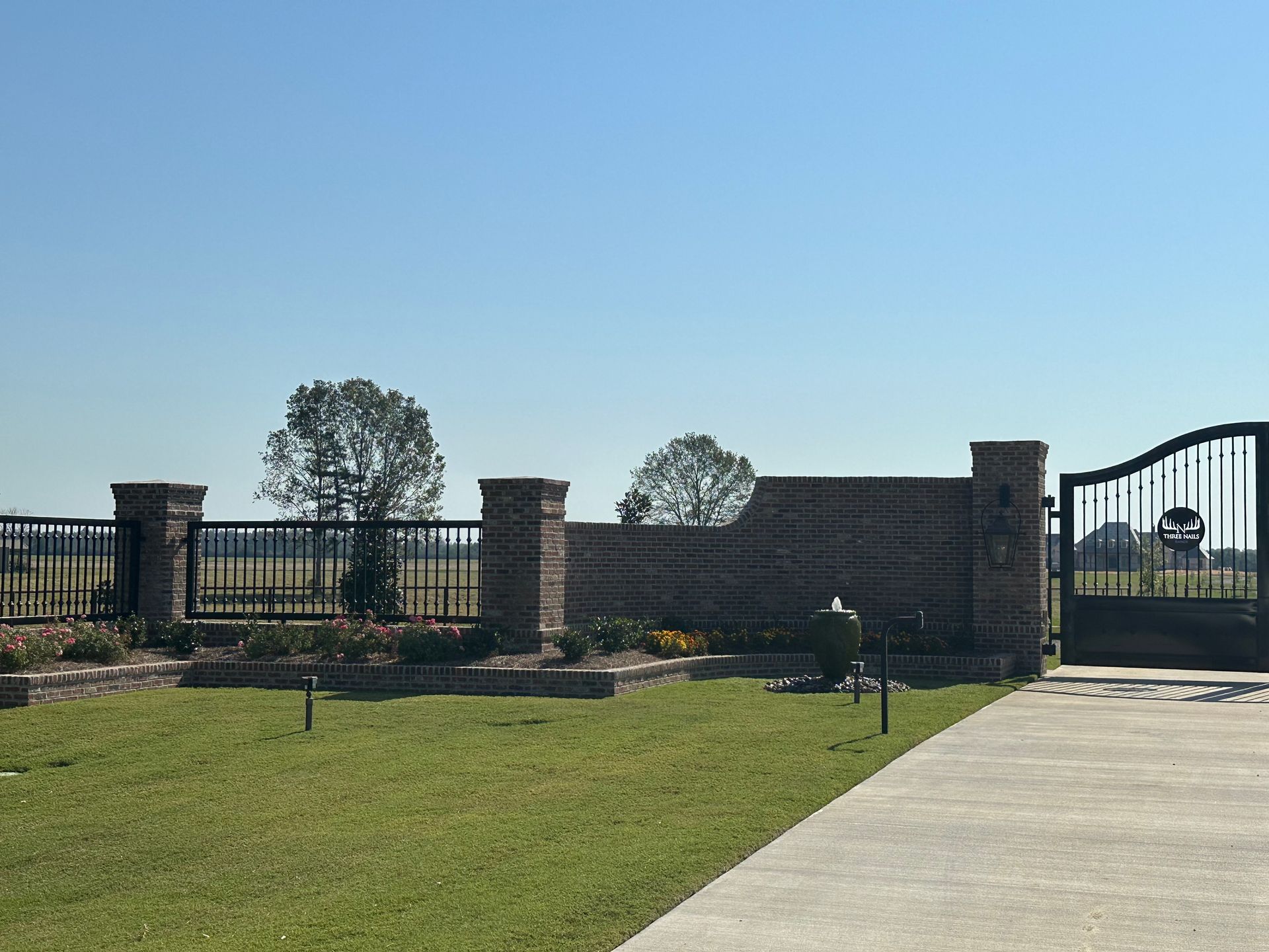 Brick entrance gate with iron fence, landscaped lawn, and a paved driveway under a clear blue sky