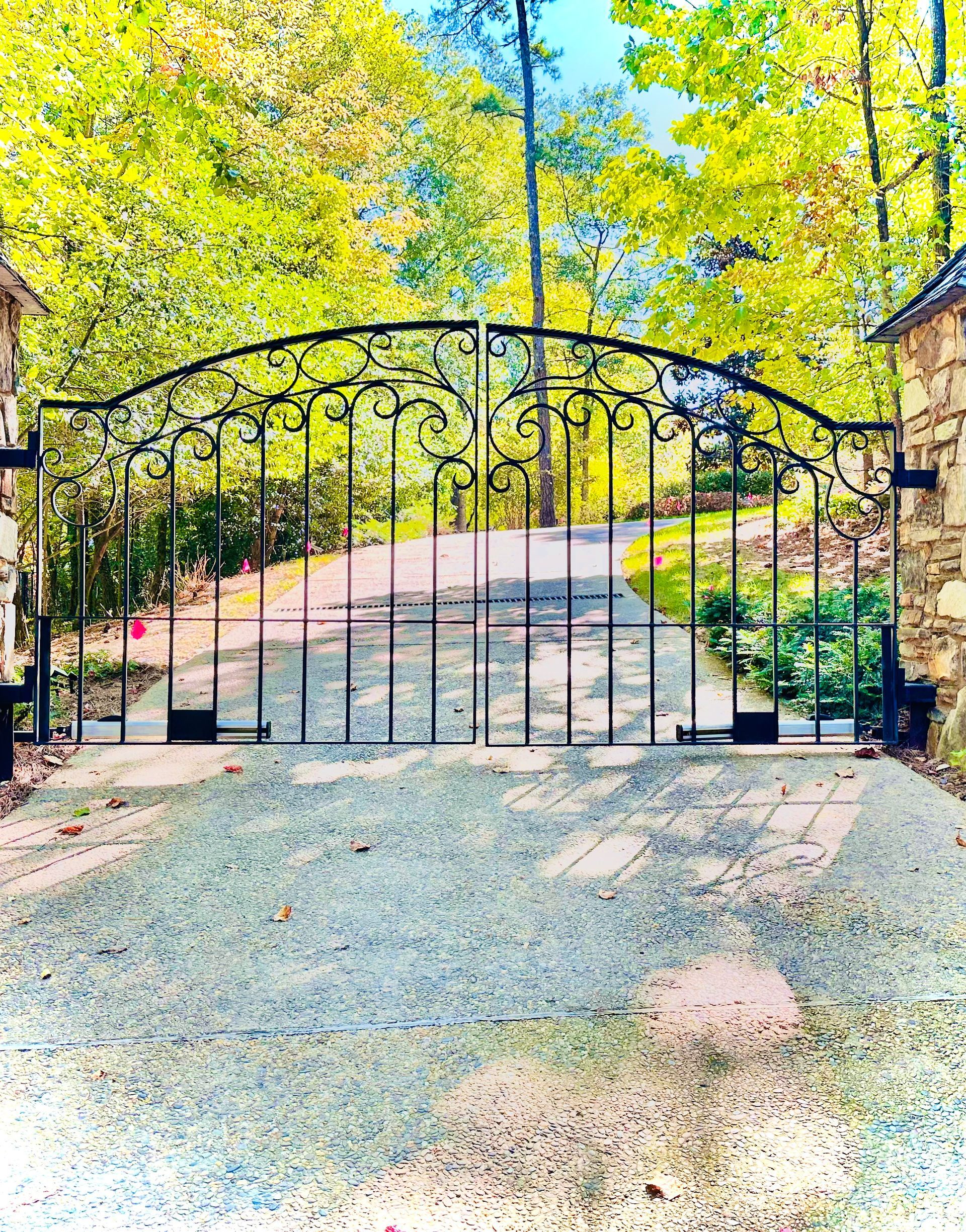 Wrought-iron gate at driveway entrance, framed by trees and autumn foliage under bright sunlight.