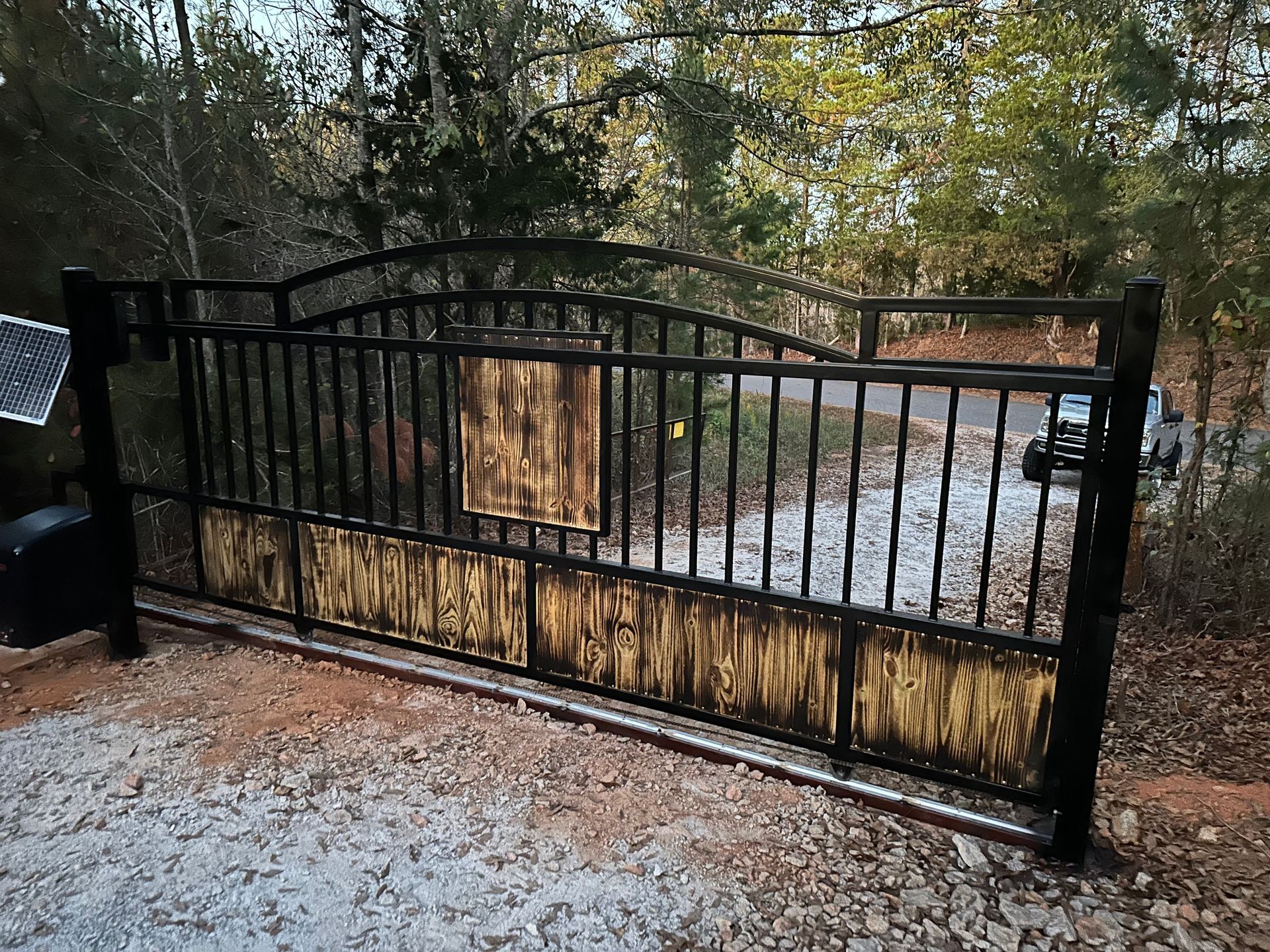 Black metal driveway gate with arched top beside a gravel driveway and wooded yard