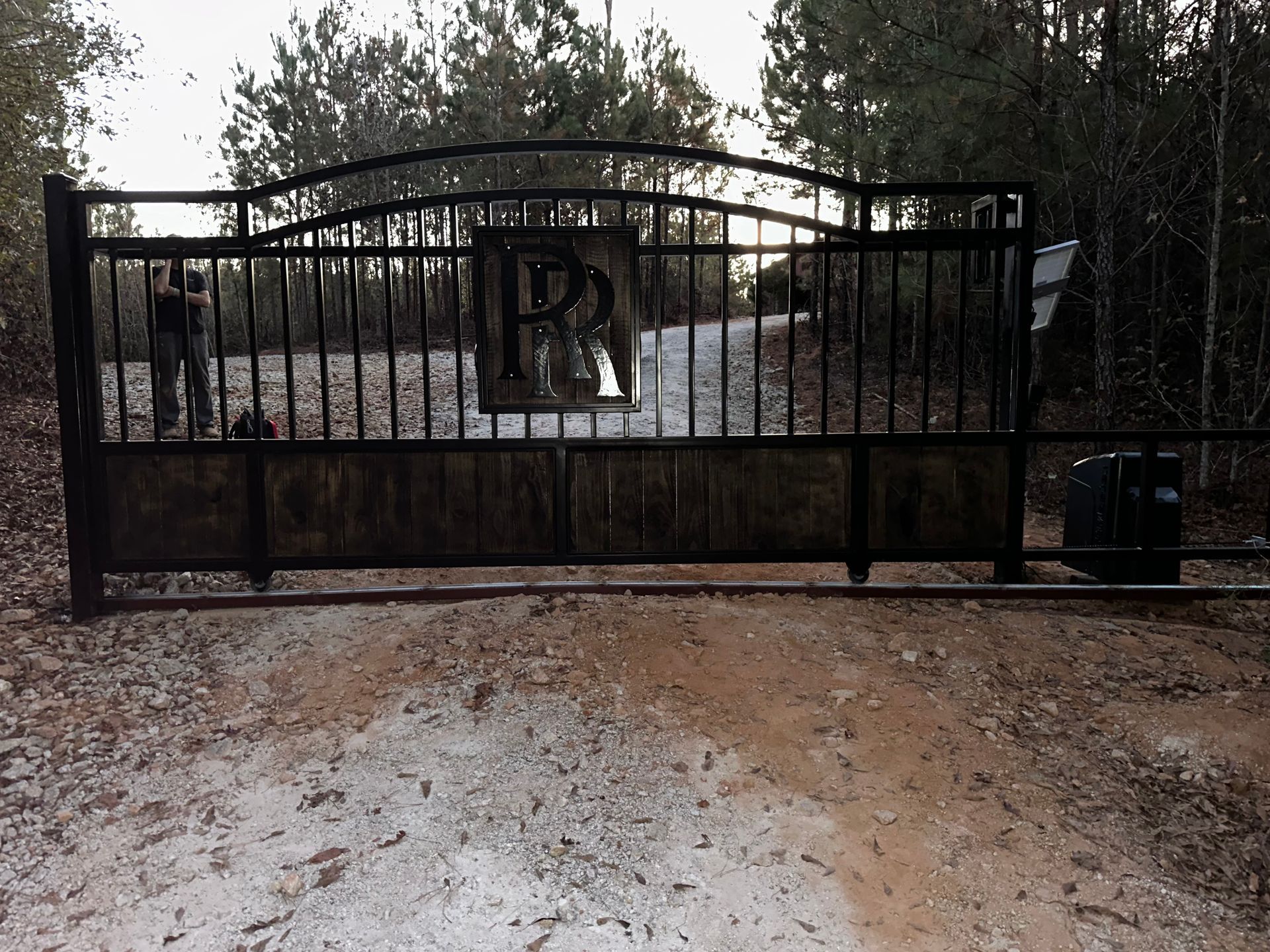Black metal gate with a large “R” across a dirt driveway, trees in the background.