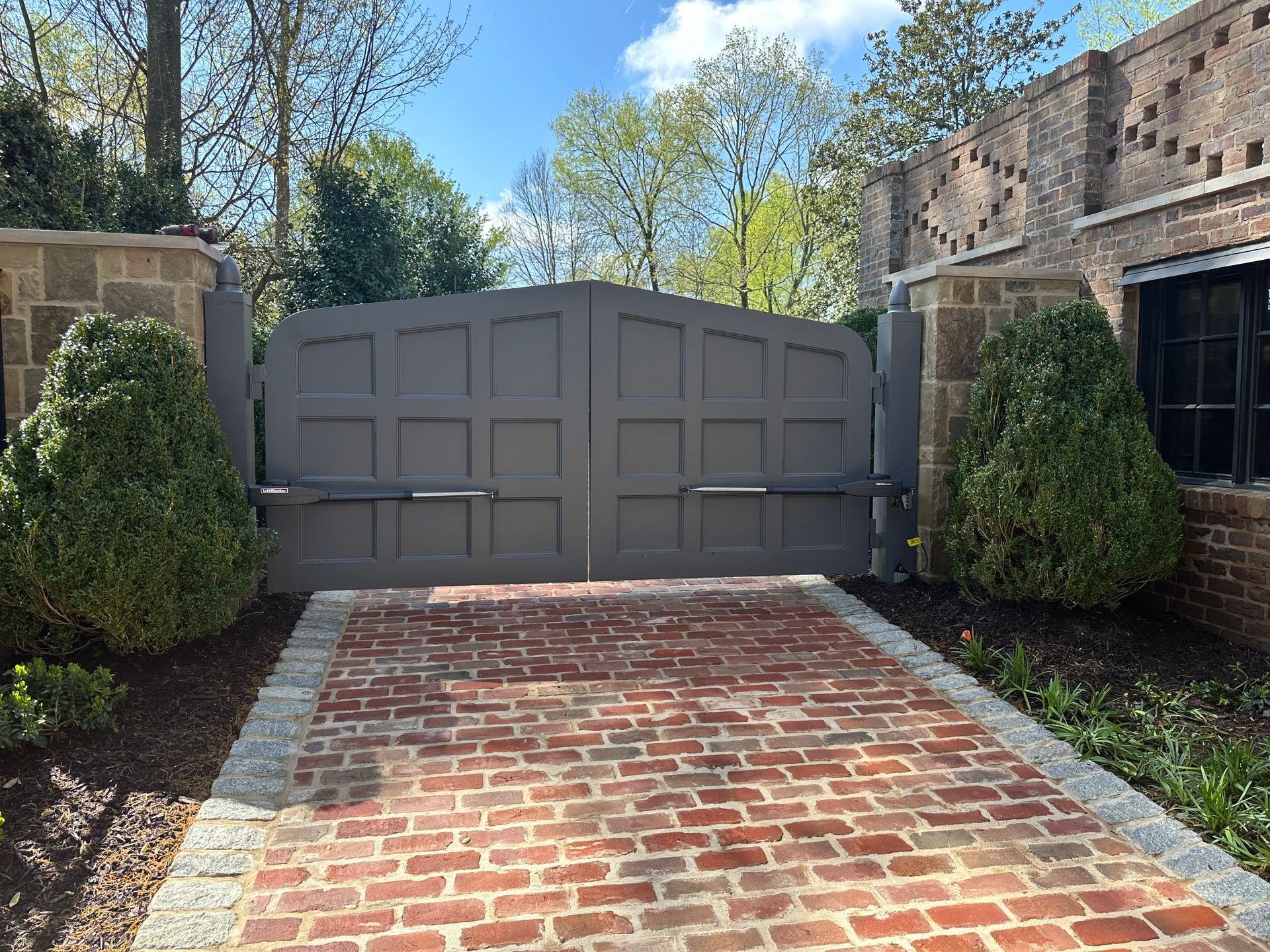 Closed gray driveway gate between stone pillars, with a brick-paved driveway and trees in the background