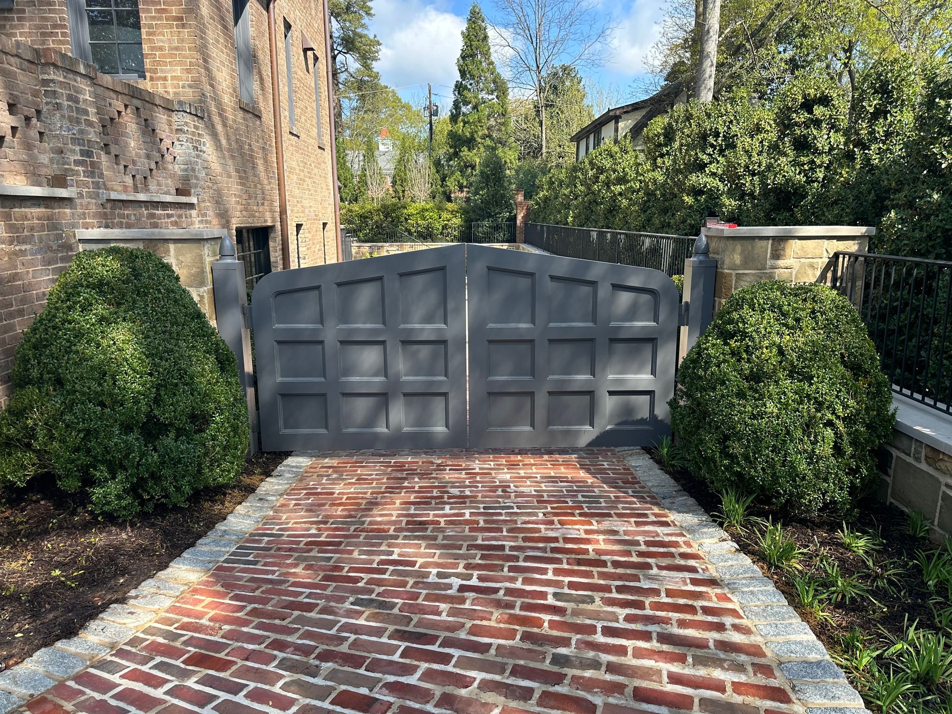 Brick driveway leading to a closed gray gate between manicured shrubs and stone walls