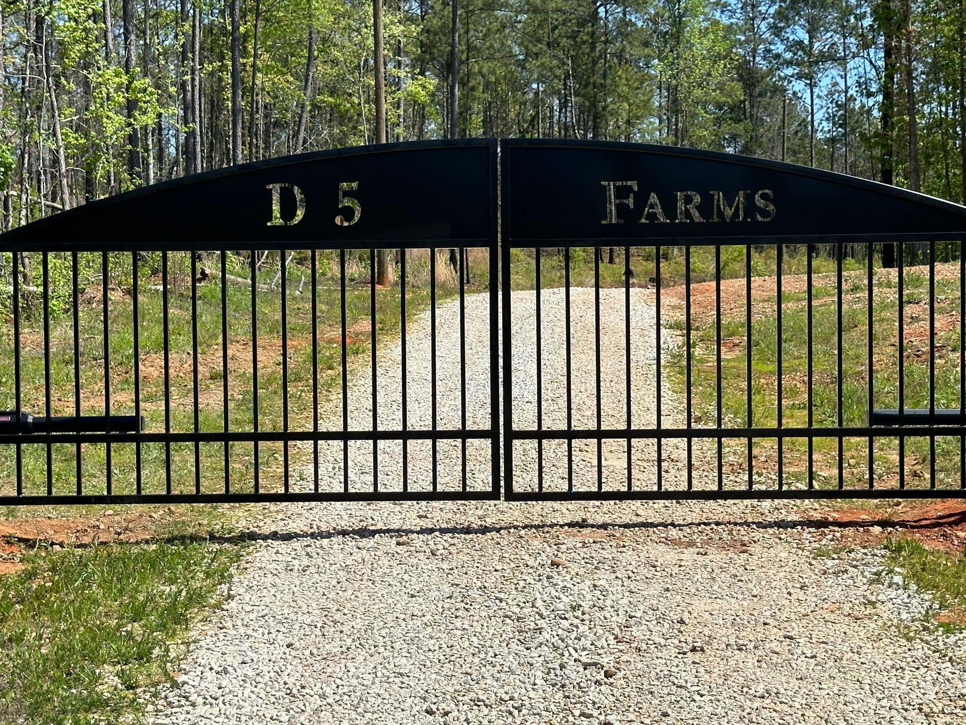 Black gate at the entrance to D5 Farms with a gravel driveway and wooded background