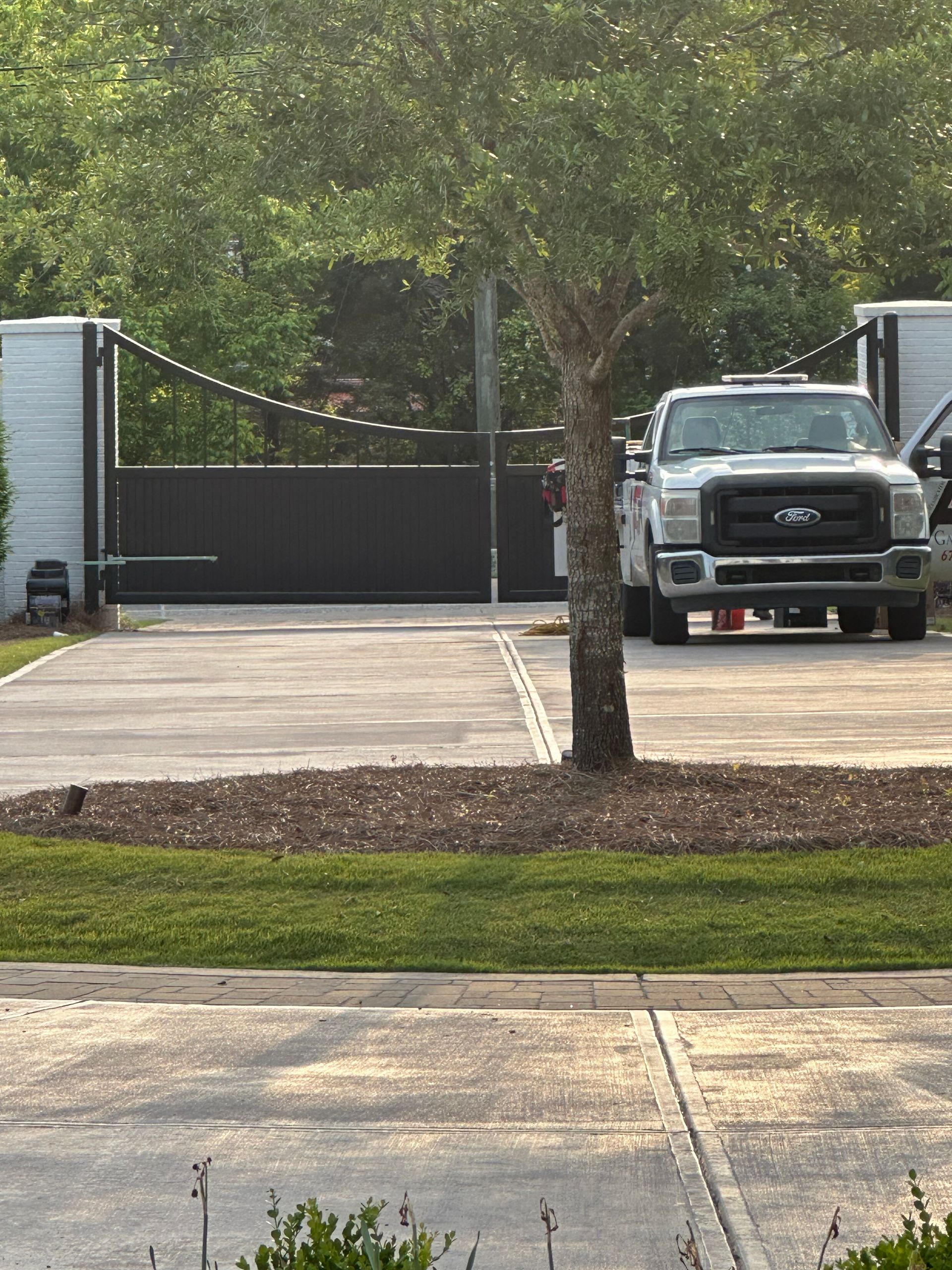 Driveway leading to a black gate with a tree in front and a parked pickup truck on the right.