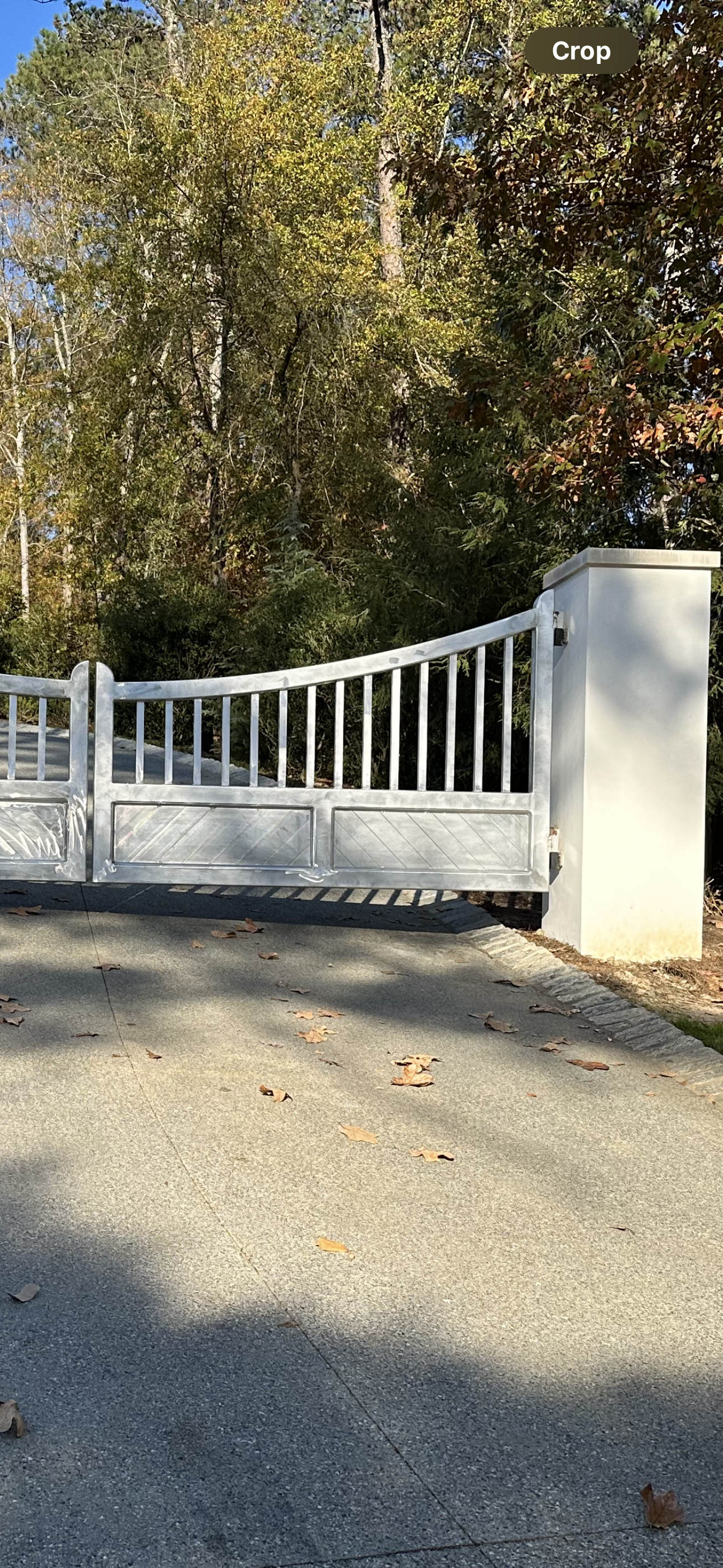 White metal gate beside a white pillar, closed across a driveway with trees behind it.