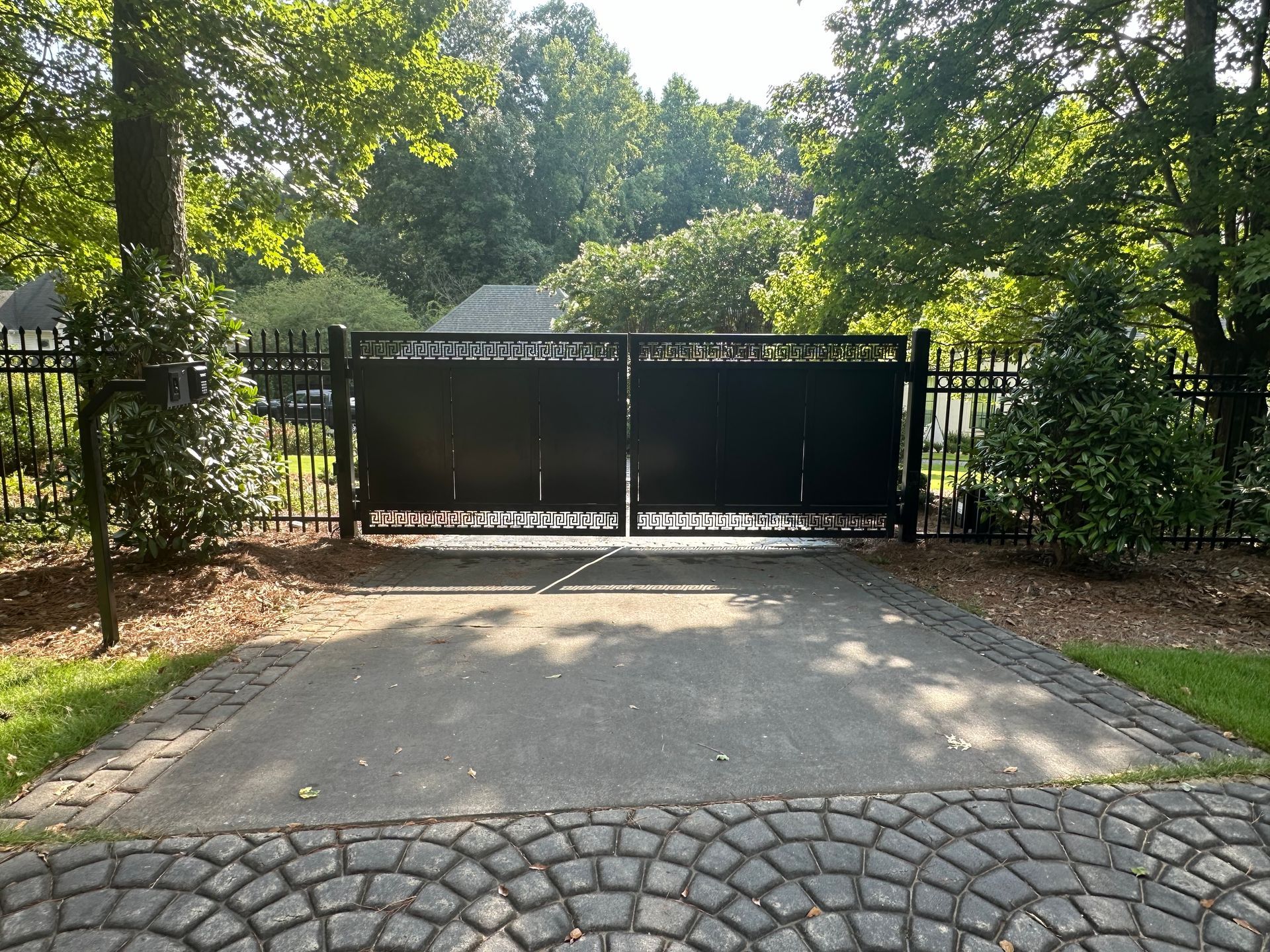 Closed black gate at the end of a driveway, surrounded by trees and greenery.