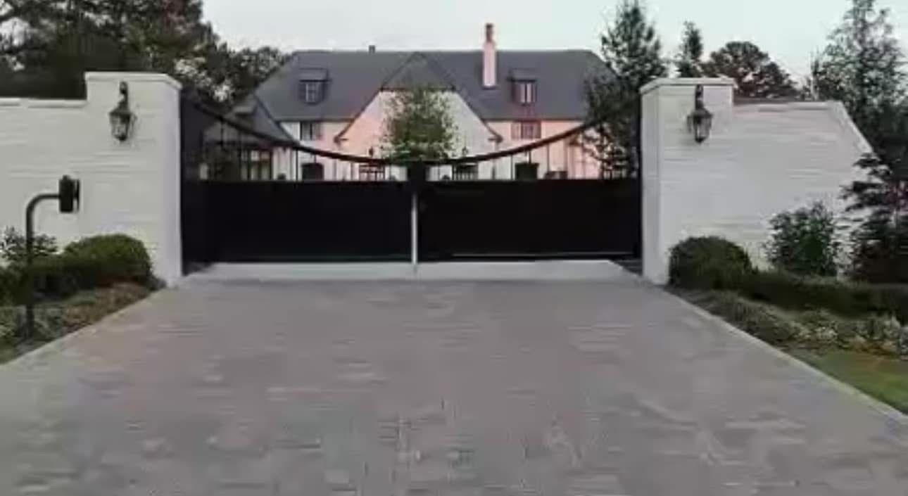 Driveway leading to a gated estate with white pillars and a large house beyond.