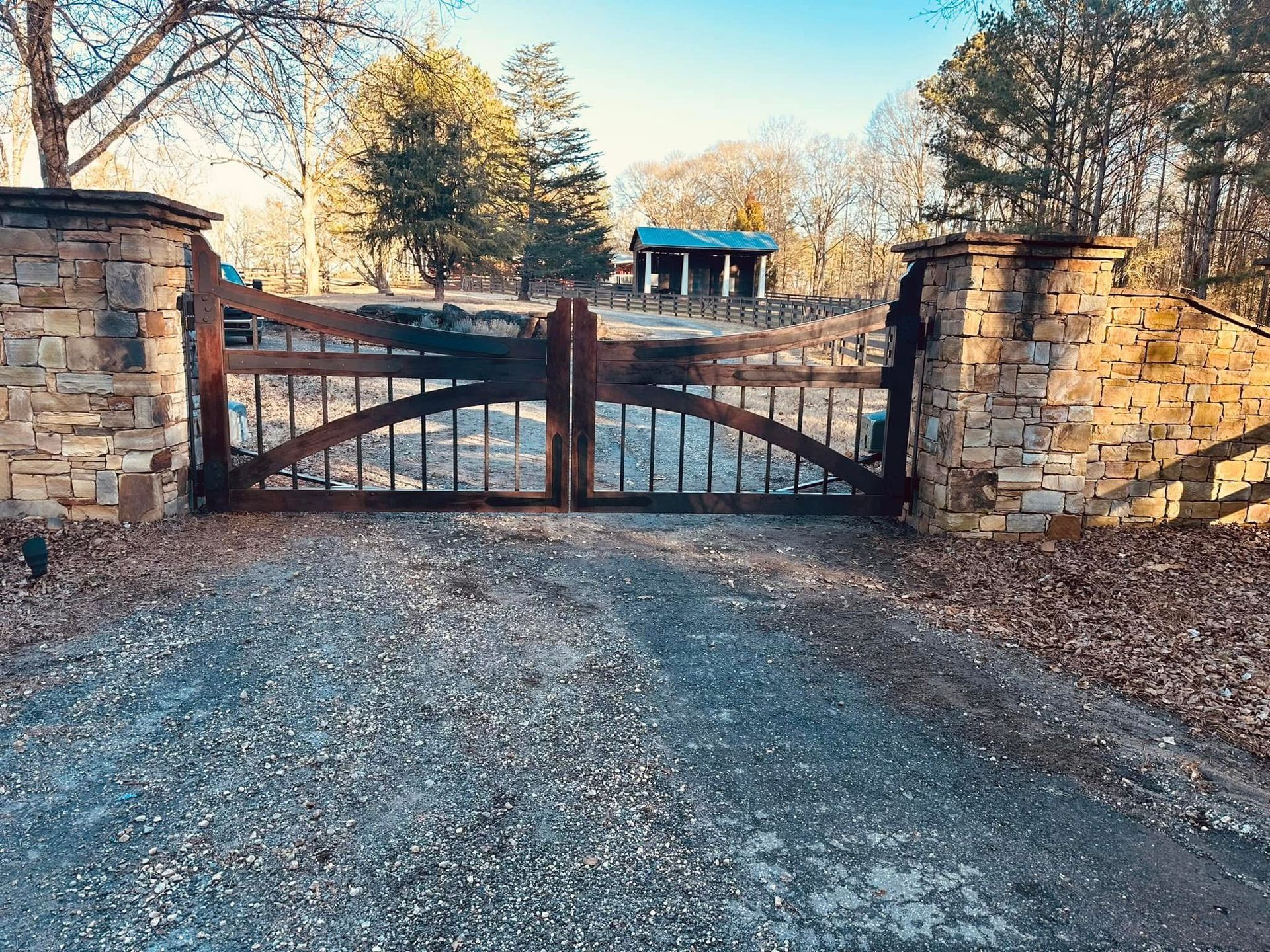 Stone-and-wood gate at the entrance to a gravel driveway, with trees and a small blue structure beyond.