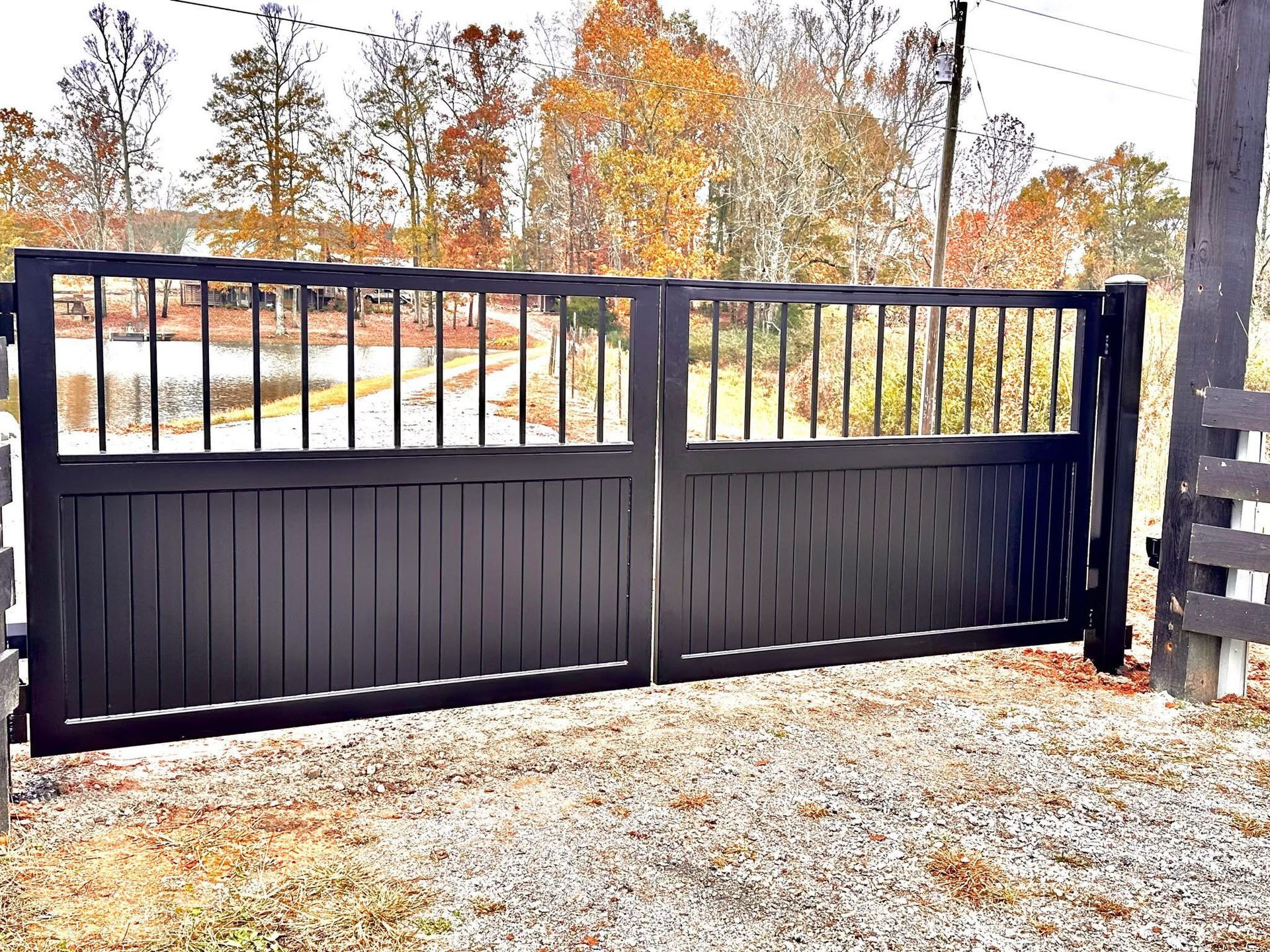 Black metal driveway gate with vertical bars and wooden posts beside a rural road in autumn
