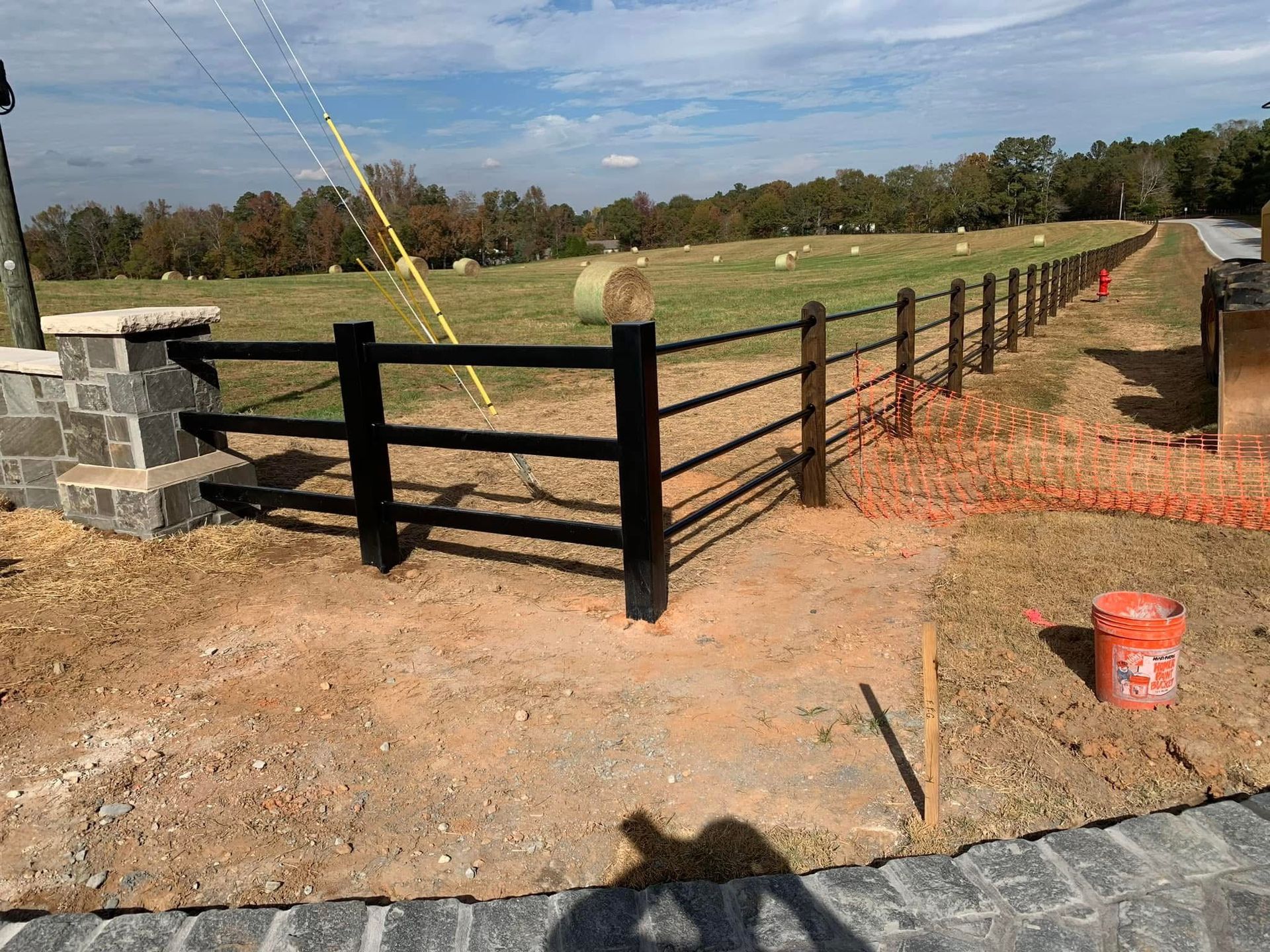 Black wooden fence around a dirt field with stone posts and a gate under a partly cloudy sky