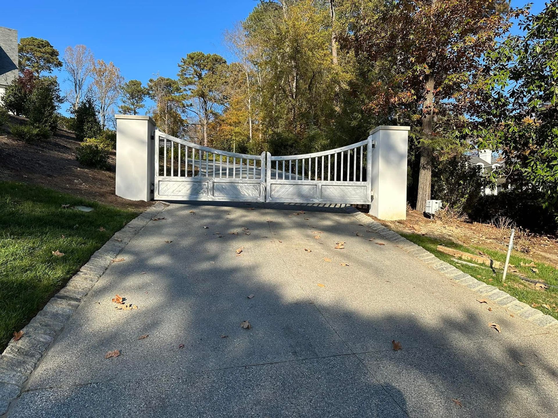 Closed white gate at the top of a sunlit gravel driveway, surrounded by trees and grass